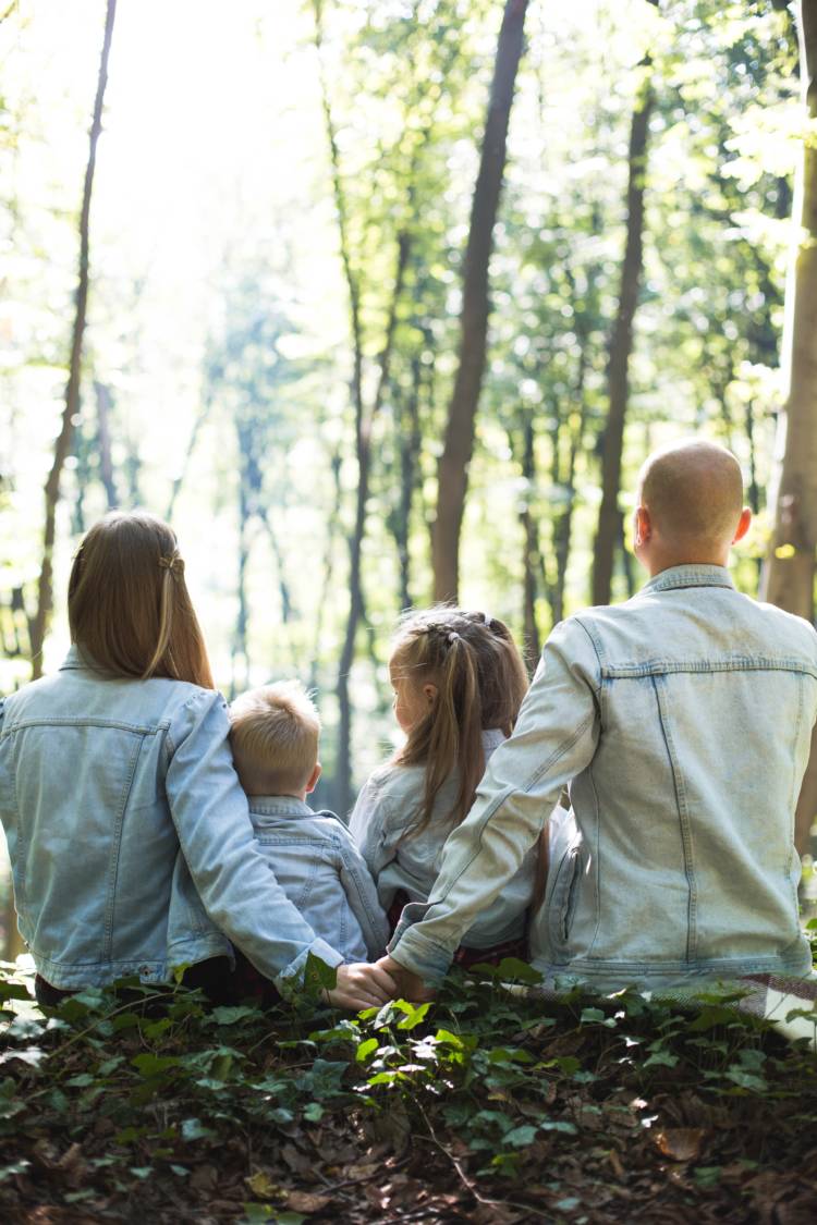 Farkkutakkeihin pukeutunut perhe istuu metsässä selin kameraan. Perheessä on äiti, isä ja 2 pientä lasta. Äiti ja isä pitävät toisiaan kädestä kiinni. 
